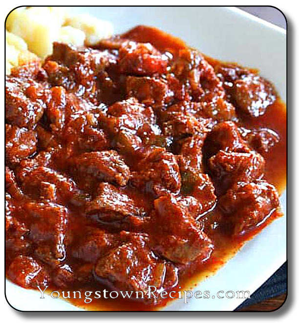 A plate of beef goulash with a rich, reddish-brown sauce, next to a side of mashed potatoes.